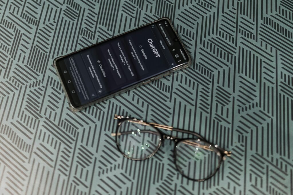 a mobile phone with ChatGPT up on the main screen rests on a bed with a pair of glasses that sits near the phone.