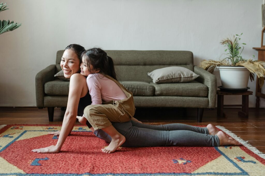 a woman is in "upward facing dog" pose on her mat at home while daughter is on her back. They are both smiling.