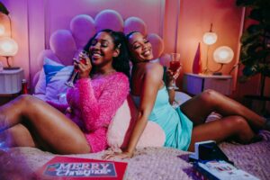 2 besties sitting back to back on a bed drinking champagne. They are laughing and smiling. They are dressed up as if they are going for a night out on the town.