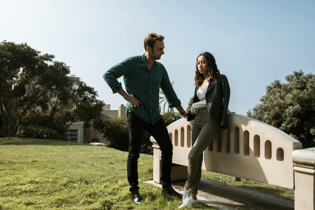 an unhappy couple are outdoors. the man is in a position of superiority as the female sits and looks down making no eye-contact