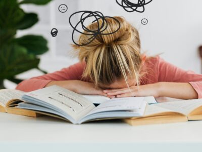 FEATURED IMAGE: get your life together - a woman lays her head down on her desk surrounded by clutter and open books in defeat