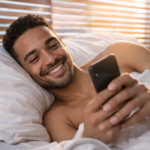 Handsome man lying on his side in bed, smiling while checking his phone in the morning as warm sunlight streams through window blinds, creating a cozy and intimate wake-up atmosphere.