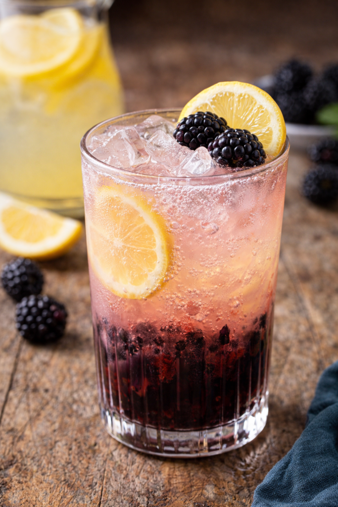 A refreshing blackberry lemonade mocktail with ice and sparkling bubbles, layered with muddled blackberries and lemonade, garnished with fresh blackberries and a lemon slice on a rustic wooden surface.