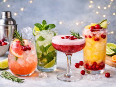 A realistic photo of four refreshing non alcoholic mocktails on ice, garnished with citrus slices, mint, rosemary, and cranberries, arranged on a light surface with fresh fruit and cocktail tools in the background.