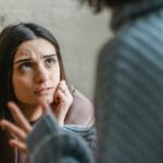 A young woman with a concerned expression rests her chin on her hand while listening to someone speaking, their hand gesturing in the foreground during an intense conversation.