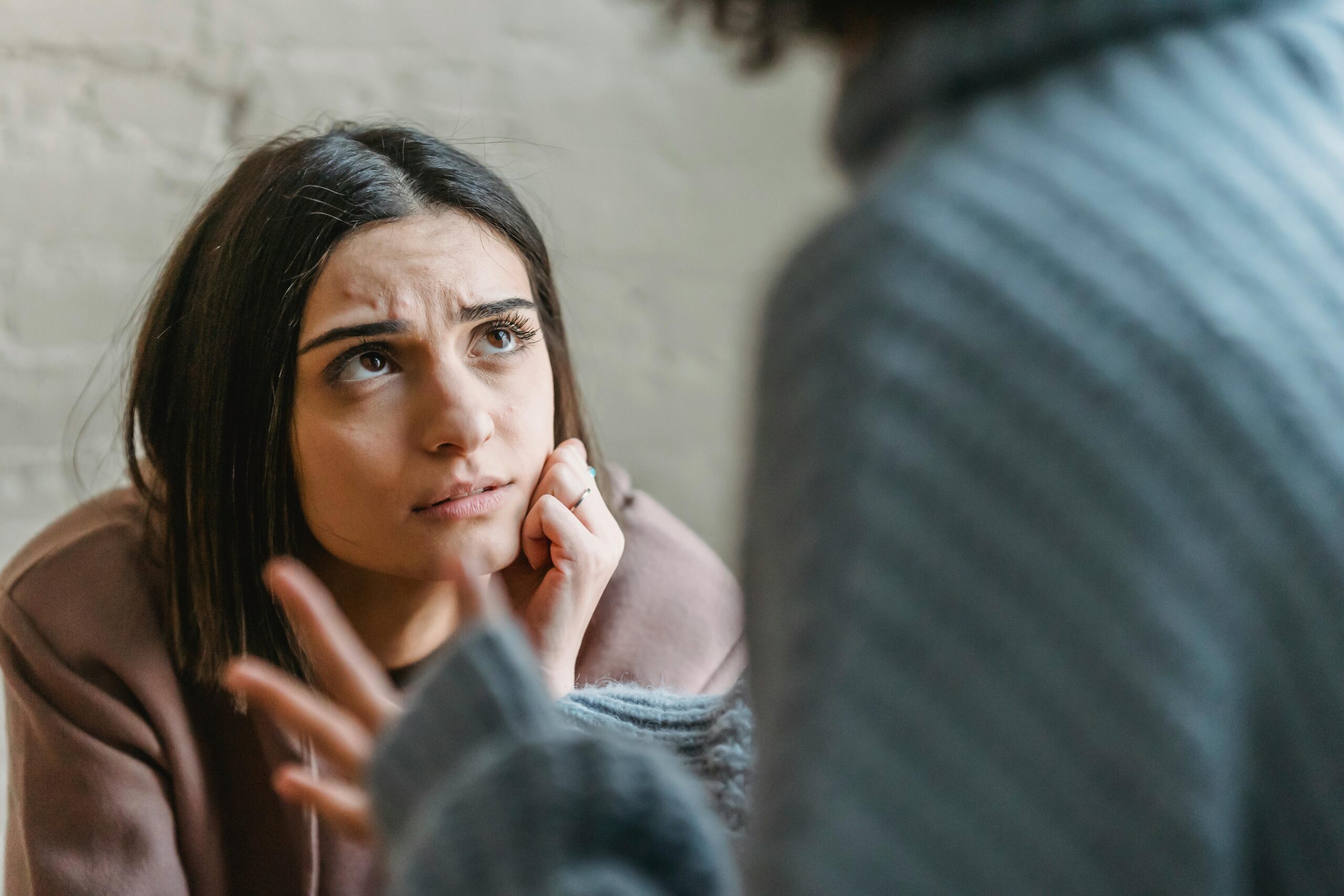 A young woman with a concerned expression rests her chin on her hand while listening to someone speaking, their hand gesturing in the foreground during an intense conversation.