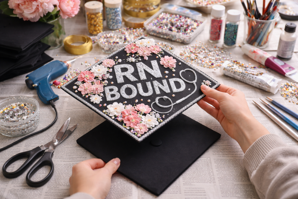 Hands placing a decorated graduation cap topper onto a plain cap on a covered workspace, surrounded by craft supplies like rhinestones, glue, scissors, and glitter.