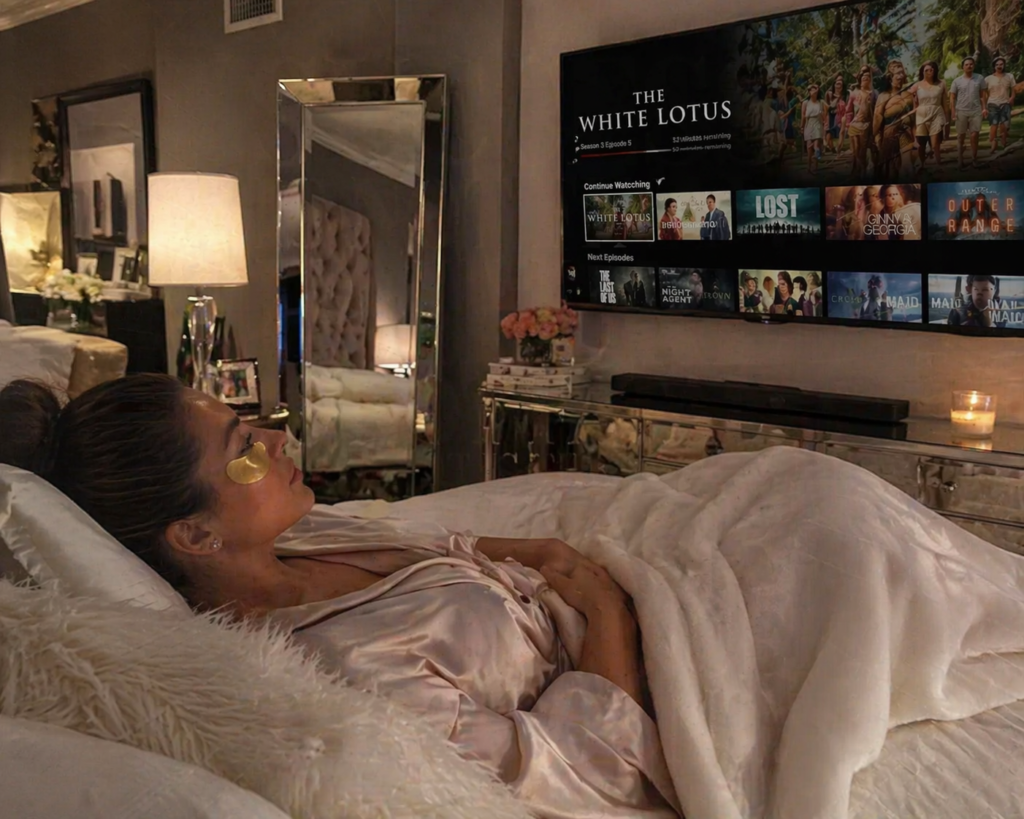 A woman lying comfortably on her bed at home wearing gold gel under-eye masks while watching a flat-screen TV. The bedroom is stylish and cozy, featuring soft lighting, decorative mirrors, neatly arranged furniture, and elegant decor, creating a relaxing self-care atmosphere.