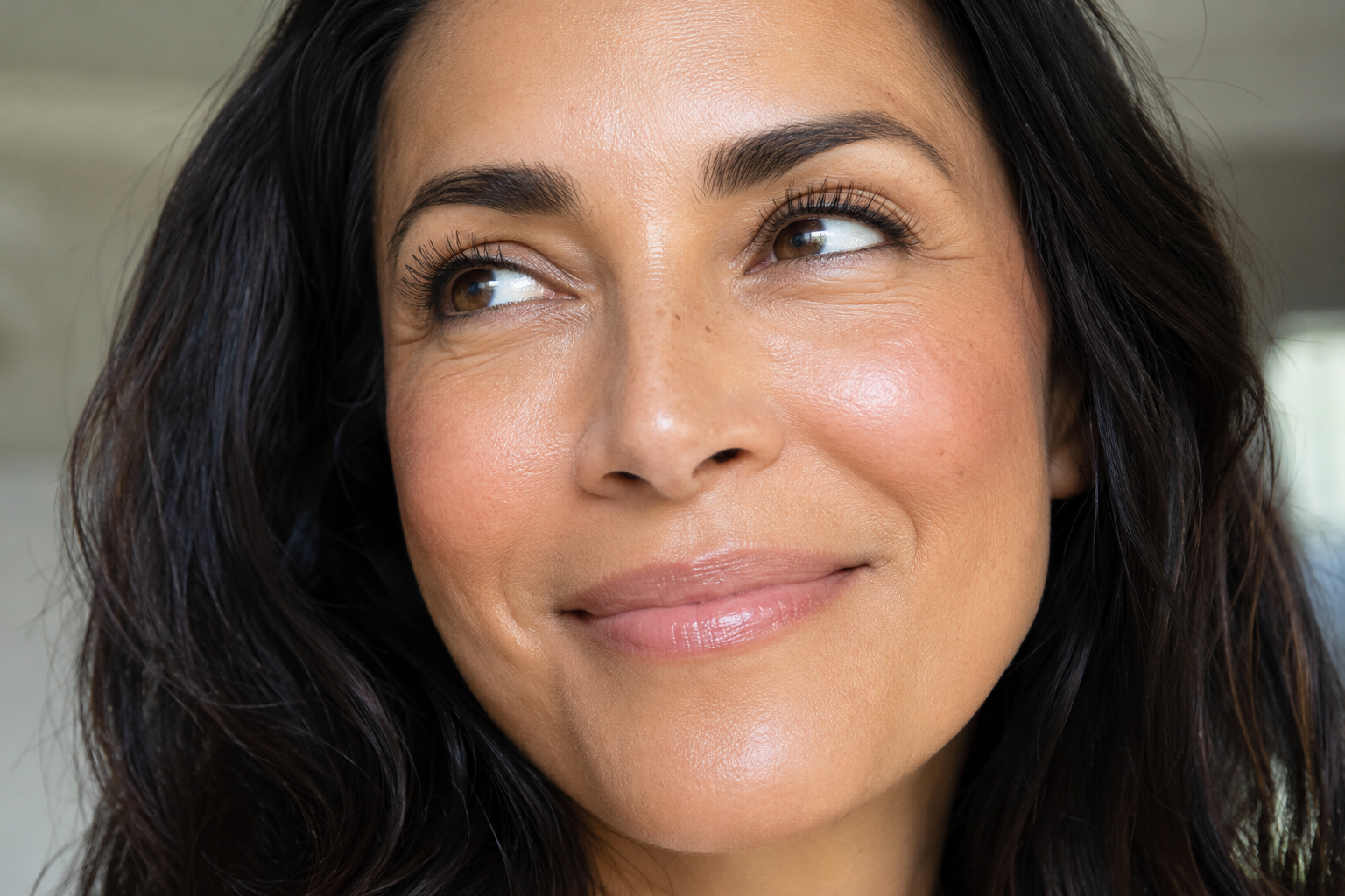 Close-up portrait of a smiling 45-year-old tan woman with healthy, glowing skin, soft cream blush, and a dewy complexion, looking upward toward the left against a softly blurred background.