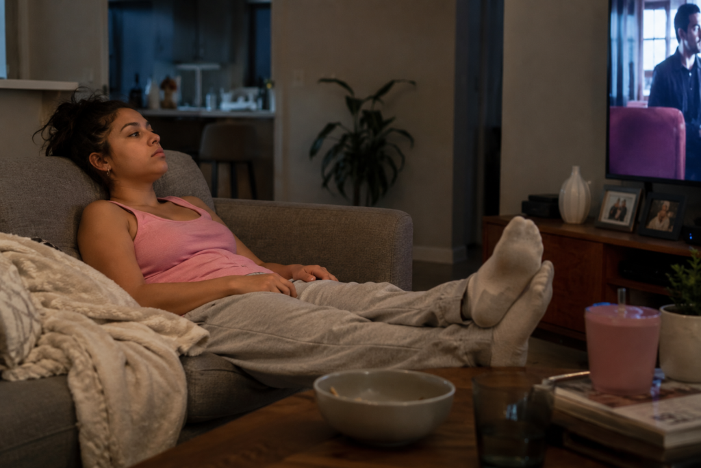 Weekend living room scene showing a woman in a pink tank top, gray sweatpants, and socks slumped on a couch with her feet propped on the coffee table. Her head rests against the back cushion, her black hair is in a messy ponytail, and she stares past the TV with a tired, disengaged expression.