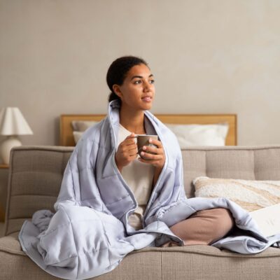 Woman wrapped in a light gray weighted blanket while sitting on a couch and holding a warm mug. A book rests beside her in a calm, neutral-toned living space. The cozy scene conveys relaxation, comfort, stress relief, and the calming benefits of a weighted blanket for rest, self-care, and emotional wellness.