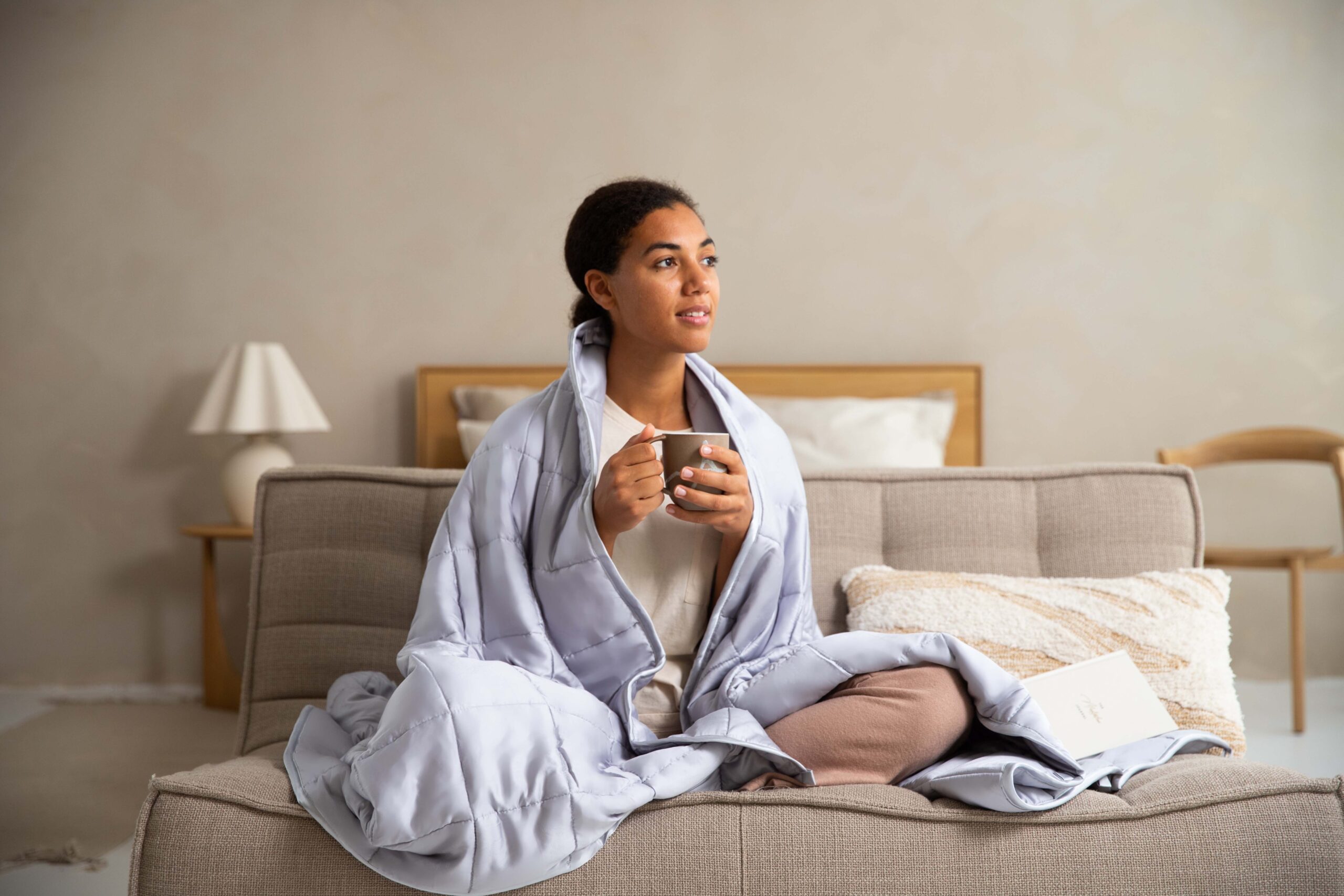 Woman wrapped in a light gray weighted blanket while sitting on a couch and holding a warm mug. A book rests beside her in a calm, neutral-toned living space. The cozy scene conveys relaxation, comfort, stress relief, and the calming benefits of a weighted blanket for rest, self-care, and emotional wellness.