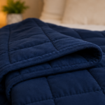 Close-up of a folded navy blue weighted blanket on a neatly made bed, showcasing its soft quilted fabric and evenly stitched pockets. A warm bedside lamp and blurred bedroom décor in the background create a calm, cozy atmosphere that represents restful sleep and relaxation.